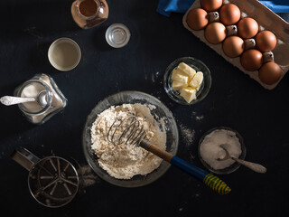 Cooking a cake. On the table are ingredients for making a pie (flour, eggs, cocoa, butter, sugar, salt), kitchen utensils and textiles. View from above. Flat lay. Dark background