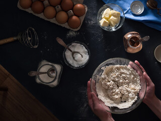 Cooking a cake. On the table are ingredients for making a pie (flour, eggs, cocoa, butter, sugar, salt), textiles. Kneading the dough. Hands in the frame. Top view of the table and floor. Flat lay. 
