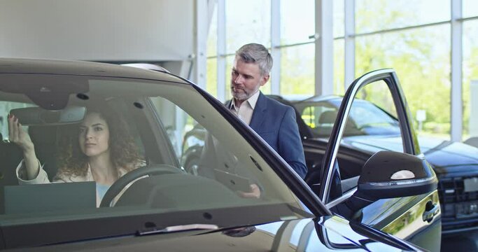 Buyer is sitting and looking around in car. Happy customer talking and listening with trader in car dealership. Auto dealership customer is standing near car, talking about specificationf of the car.