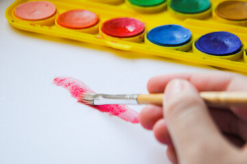 Close up on a hand holding a brush and painting on a white sheet a red pattern with a watercolor...