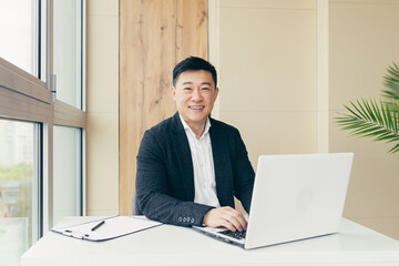 Successful Asian businessman, in a black business suit, works on a laptop, relaxes in a stylish office