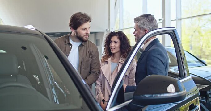 Confident caucasian male trader with tablet opens car door for the customers and talking about specifications of the car Young couple choosing new automobile. Car dealership, car business.