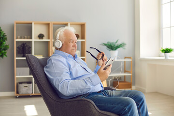 Senior man sitting in armchair at home wearing headphones and using mobile phone