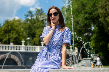 Young woman in dress and glasses sits near a fountain in the park