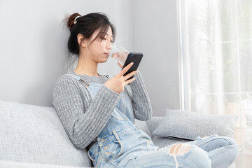Asian girl sitting on the sofa drinking water and looking at the mobile phone