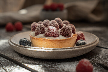 Delicious tartlet with custard, fresh raspberries and sugar powder, served on gray ceramic plate over wooden surface. Summer seasonal sweets. Soft focus.