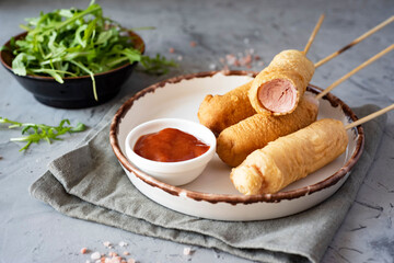 Quick snack: corn dogs on sticks. Sausages in batter on sticks on a white plate on a gray background.