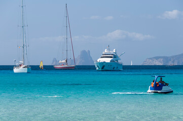 Fototapeta premium A couple of tourists sail near the beach in Formentera with a small boat. In the background luxury boats anchored in the Mediterranean Sea.