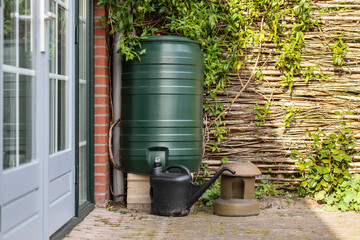 A green rain barrel to collect rainwater and reusing it to water the paints and flowers in a backyard with a wattle fence made of willow branches on a sunny day - obrazy, fototapety, plakaty