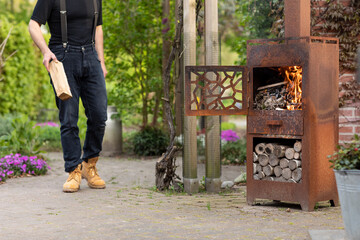 A man carrying a wooden log to a metal wood burner, burning wood stove in his garden while living...