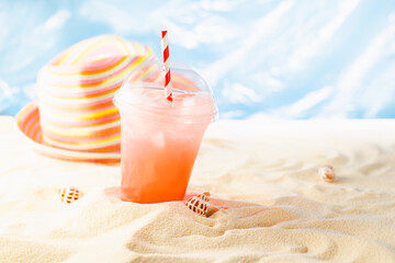 Summer vacation on seaside coast on sunny beach with ocean view with fresh cold pink fruit cocktail with straw in transparent takeaway mug and sun hat.
