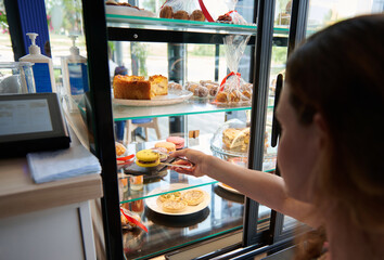 Pastry cafe worker selects delicious yellow macaron from a showcase with a large assortment of pastries and sweet desserts