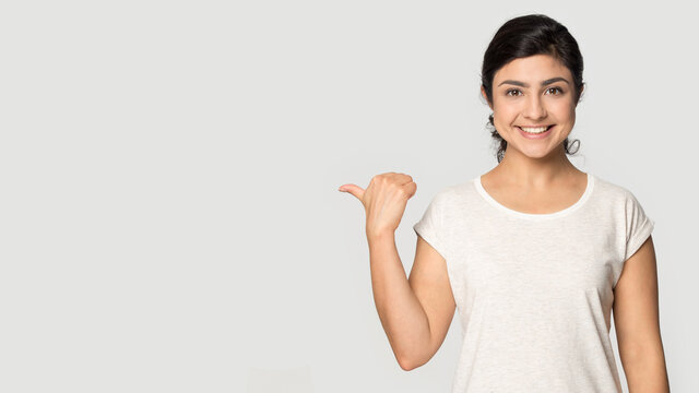 Portrait of smiling Indian woman isolated on grey studio background point at blank empty copy space. Happy millennial mixed race female show good promotion deal or sale discount. Commerce concept.