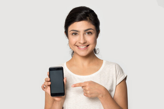 Headshot portrait of smiling young Indian woman isolated on grey studio background point at cellphone screen good online deal. Picture of happy mixed race female user show promotion on smartphone.