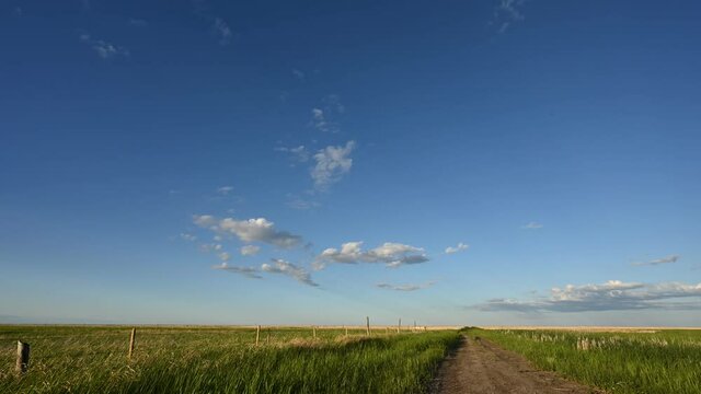 Early evening clouds drift through a blue sky in this time lapse over a prairie setting with a dirt road and fences.
