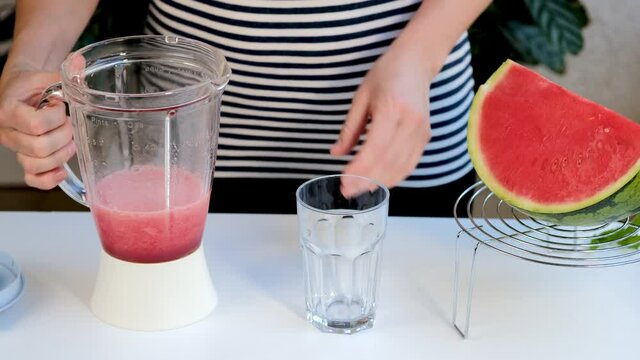 White woman pours glass of watermelon smoothie from a blender. Recipe of fresh healthy drink. Girl cooking sweet fruit smoothie. Healthy summer eating concept.