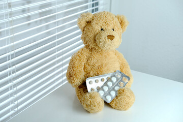 teddy bear, medications, white pills in a blister, against the background of blinds on the table, concept getting sick, having a cold, skipping school, kindergarten