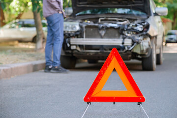 Red triangle warning sign of car accident on road in front of wrecked car. Man inspecting wrecked...