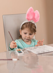 A little girl in cute headband with pink bunny ears draws with a brush with watercolor paints