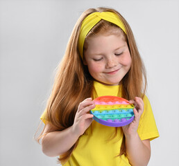 Cute little red-haired girl with colorful pop simple silicone toy with dimples shows surprised, looks at the toy. Studio face portrait children shot