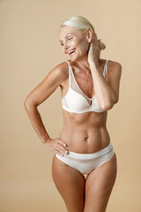 Fototapeta premium Charming mature woman in underwear with fit perfect body smiling and touching her head, standing with eyes closed isolated over beige background