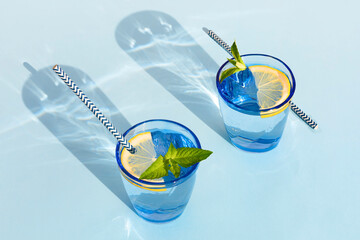 summer drink. cool refreshing beverage with mint and lemon in colored glass with paper tube. zero waste home. minimalism. natural fresh lemonade with hard shadows on blue background - obrazy, fototapety, plakaty