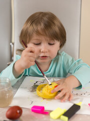 A three-year-old girl in a turquoise blouse enthusiastically paints yellow chicken with paints and a marker