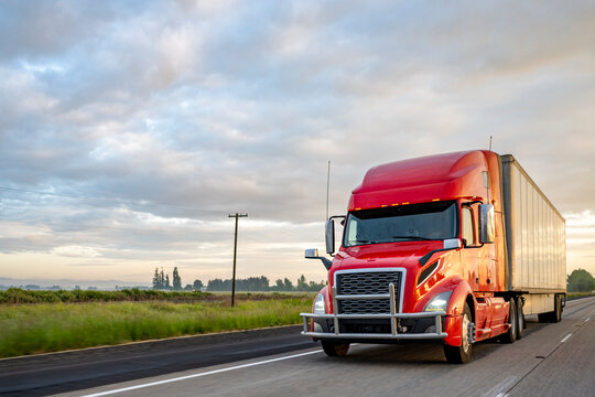 Bright red big rig bonnet semi truck transporting cargo in dry van semi trailer running on the evening highway road