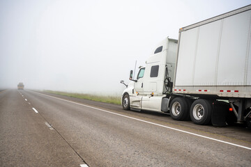 Broken white big rig semi truck with dry van semi trailer stands on the side of a foggy road...