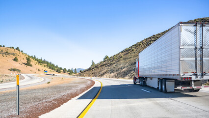 Classic red powerful industrial big rig semi truck transporting frozen food in refrigerator semi trailer running on the winding highway road between the hills