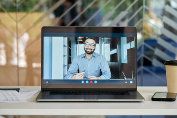 Laptop computer at office workplace table with smiling indian businessman executive leader, coach on pc screen having video chat, conference call, virtual meeting or giving training on videocall.