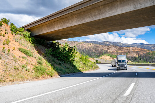 Convoy Of Two Big Rig Semi Trucks With Semi Trailers Running On The Turning Highway Road With Concrete Bridge Across