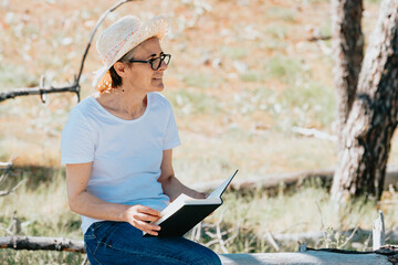 Old woman reading a book at the beach during a super sunny day. Freedom concept with copy space, relaxation and senior retirement happiness