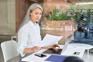 Serious focused Asian older business woman ceo manager sitting at desk doing financial paperwork using pc laptop making online tax, loans, debts bank payments in contemporary corporation office.