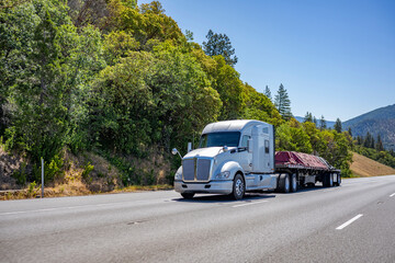 Powerful white big rig semi truck transporting tarped cargo on flat bed semi trailer climbing uphill on the wide mountain road