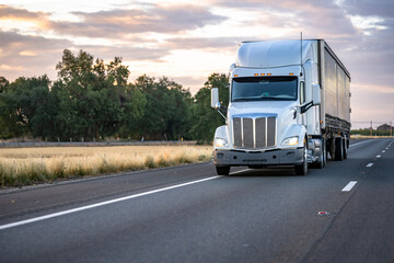 Powerful white big rig semi truck with turned on headlight transporting cargo in dry van semi...