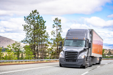 Shiny gray big rig semi truck with orange dry van semi trailer moving on the road for cargo delivery