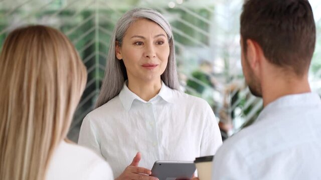 Asian senior businesswoman ceo sales agent mentor talking to young Caucasian male and female interns trainees discussing business project using tablet device standing in modern corporate office.