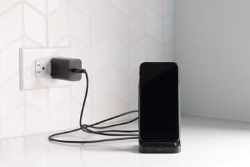 a black smart phone docked in a wireless charging station plugged in on a white kitchen counter top or bench with copy space