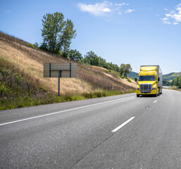Obraz premium Well-remembered bright yellow big rig semi truck transporting loaded semi trailer running on the highway road between hills