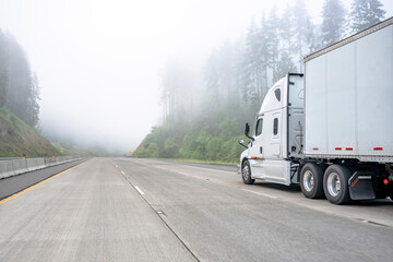 White big rig semi truck with dry van semi trailer running on the dangerous foggy highway road with poor visibility