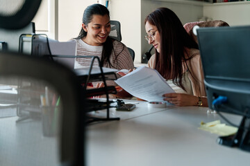 two young latina business women talking and working together in office, using documents and office supplies, teamwork concept