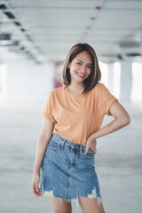 Stylish Asian girl wearing orange t-shirt and jean skirt posing in empty room, urban clothing style. Street photography.