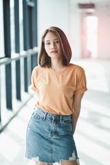 Stylish Asian girl wearing orange t-shirt and jean skirt posing in empty room, urban clothing style. Street photography.