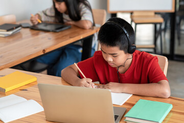 Asian man student wearing headphones sitting in taking notes with laptop in class room.