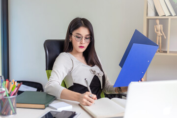 Young Asian businesswoman wearing glasses holding document and pen taking notes while serious working at the office.