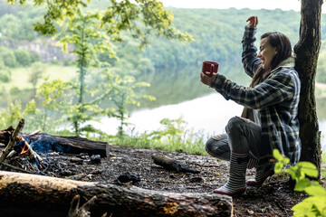 A girl with a cup in her hands enjoys nature near a fire in the forest.