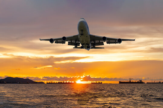 front image commercial passenger aircraft or cargo airplane fly over coast of sea in evening with golden sunset seascape view and bulk cargo ships anchored at horizon line