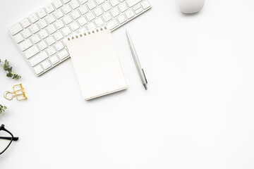 Small blank notebook with pen are on top of computer keyboard over white woman office desk table with office supplies. Top view with copy space, flat lay.