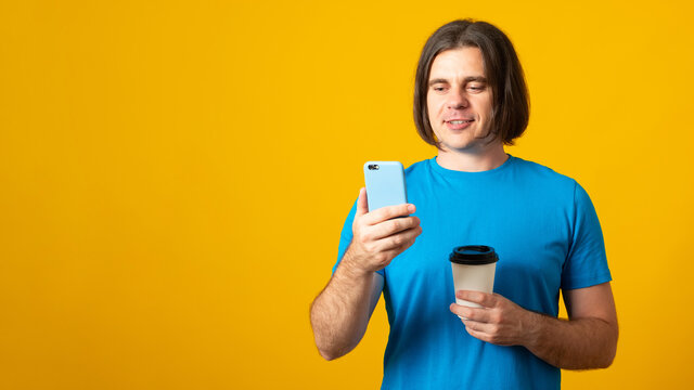 Banner. Happy handsome man uses mobile phone for online communication and surfing internet holds takeaway coffee being in good mood dressed casually poses over vivid yellow wall.